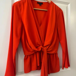 Röd omlottblus med knyt från Topshop - Snygg röd omlottblus från Topshop med v-ringning, långa ärmar och volangkant nedtill. Blusen har ett brett knytband i midjan som ger en markerad siluett. Perfekt för dig som vill sticka ut med en färgstark och trendig topp.