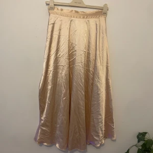 Champagnefärgad satinkjol med knyt - Snygg långkjol i skimrande champagnefärgad satin med knytband i midjan. Kjolen har en mjuk, flowy passform och ger en lyxig känsla med sitt glansiga material. Perfekt för dig som vill sticka ut med en elegant och trendig look.