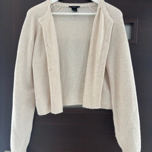 Beige kofta från Lindex  - Supermysig beige kofta från Lindex i stickad kvalitet. Koftan har långa ärmar, ribbade kanter och knappar framtill. Perfekt att slänga över en t-shirt eller linne när det blir lite kyligt. Enkel och stilren design som funkar till allt. Tröjan är lite nopprig. 