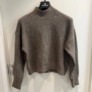 Brun stickad polotröja - Mysig brun stickad polotröja med hög krage och lång ärm. Tröjan har en loose passform och är perfekt för kyliga dagar. Den neutrala färgen gör den enkel att matcha med olika outfits. Har ett hål på baksidan som man finner på bild nr 2, men inget man lägger märke till