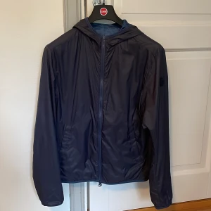 Blå reversiable vindjacka med huva från Colmar - Snygg marinblå vindjacka från Colmar med huva och dragkedja framtill. Har inte använt den så mycket så skicket är 10/10 Inga fel med den eller fläckar, den är som ny. Köpt från Zalando för 3200kr och den sitter som Small. Den är reversiable så den går att vända ut och in. Mörkblå och ljusblå camo färg på dom 2 olika sidorna. Den här en colmar loga på sidan vid vänstra armen.