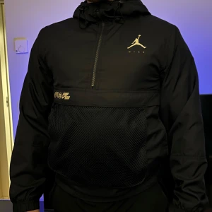 Svart Nike Jordan vindjacka med huva - Svart vindjacka från Nike Jordan med huva och halv dragkedja. Jackan har meshficka framtill,  justerbar dragsko i halsen. Perfekt för dig som gillar sportig och clean stil.  Xs men känns som S