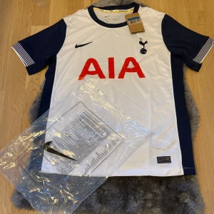 Tottenham Hotspur hemmatröja Nike - Snygg Tottenham Hotspur hemmatröja från Nike med klassisk vit front, mörkblå ärmar och röda AIA-loggan på bröstet. Tröjan har klubbmärket broderat och är tillverkad i lätt, ventilerande material. Perfekt för dig som älskar fotboll och Spurs.