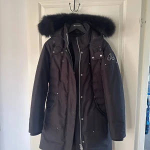 Svart dunjacka från Moose Knuckles - Säljer en svart dunjacka från Moose Knuckles med fluffig pälskant på huvan och snygga detaljer i silver. Jackan har dragkedja, knappar och logga på ärmen. Perfekt för kalla dagar med sin långa modell och vadderade partier.