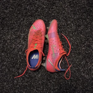 Nike mercurial skruvdubb  - Säljer ett par röda Nike fotbollsskor med gröna och silvriga detaljer. Skorna har snörning och platt sula med dobbar för bra grepp på planen. Perfekta för dig som vill sticka ut på matchen!