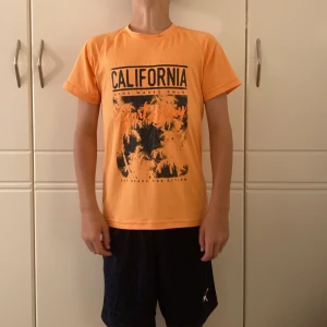 Orange t-shirt från Color Kids - Snygg orange t-shirt från Color Kids med tryck framtill där det står 'California' och palmmotiv. T-shirten har rund hals, korta ärmar och är gjord i mjukt syntetmaterial. Aldrig kommit till användning. Ny kostar 239kr