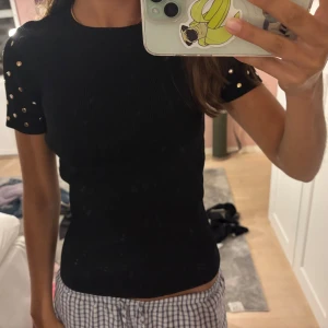 Svart ribbad topp med nitar - Svart ribbad topp med korta ärmar och coola silvriga nitar på axlarna. Tight passform som framhäver figuren och ger en edgy vibe. Perfekt att styla med jeans eller kjol för en trendig look.