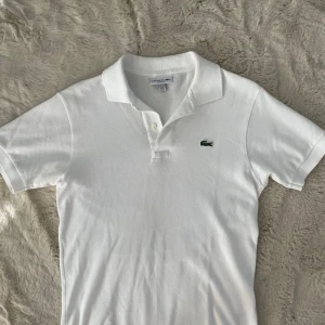Vit pikétröja från Lacoste  - Klassisk vit pikétröja från Lacoste i storlek XS. Tröjan har korta ärmar, och den ikoniska gröna krokodilloggan broderad på bröstet. Kragen saknar en av  knapparna! Tillverkad i bomull med en skön och luftig känsla. Perfekt för en clean och sportig stil.