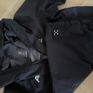 Svart vindjacka från Haglöfs - Svart vindjacka från Haglöfs med diskret logga på bröstet och dragkedja framtill. Jackan har hög krage, två stora fickor och är tillverkad i slitstark polyester. Perfekt för blåsiga dagar och outdoor-äventyr.