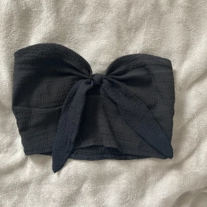 Svart bandeau tubtopp med rosett  - Svart bandeau tubtopp från SHEIN i storlek XS. Toppen är i ett strukturerat bomullstyg och har en stor knytrosett framtill som ger en snygg statement-look. Perfekt för att lyfta vilken outfit som helst med sin coola och trendiga design.
