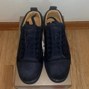 Christian Louboutin svarta sneakers mocka - Snygga svarta sneakers från Christian Louboutin i mocka med klassisk röd sula och broderad logga baktill. Skorna har rund tå, platt sula och svarta snören. Perfekta för dig som vill sticka ut med exklusiv streetstyle.