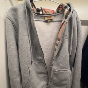Grå Burberry hoodie med dragkedja - Säljer en grå hoodie från Burberry med klassiskt rutigt foder i huvan. Tröjan har dragkedja framtill, två fickor och snörning i huvan. Perfekt för dig som vill ha en stilren och lyxig vardagslook med ikoniska Burberry-detaljer. Säljer pga att den inte passar mig längre 