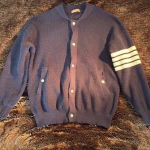 Mörkblå kofta med vita ränder - Mörkblå stickad kofta från Fashion med vita knappar och fyra vita ränder på ena ärmen. Koftan har två fickor framtill och ribbade muddar vid ärmslut och nederkant. Perfekt för lager-på-lager och snygg till jeans. Modellen är 179 65kg så skullle säga att det är en S/M