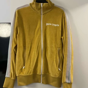 Gul track jacket från Palm Angels - Gul track jacket från Palm Angels med vita och grå ränder längs ärmarna. Tröjan har hög krage, hel dragkedja framtill och två fickor med dragkedja. Materialet är en mix av bomull och polyester för en mjuk och sportig känsla. Ikonisk logga på bröstet.