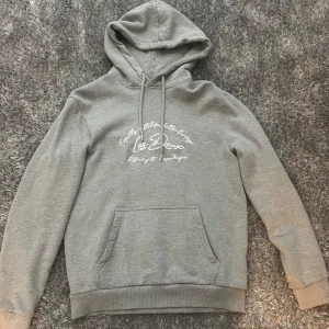 Grå hoodie från Les Deux med tryck - Säljer en grå hoodie från Les Deux med vit text på bröstet som helt oanvänd. Tröjan har huva med snörning, magficka och mjukt fleecefoder på insidan. Perfekt för kyliga dagar och riktigt skön att ha på sig. 