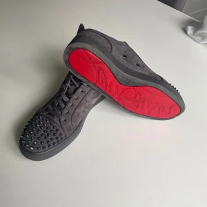 Mörkgråa louboutine med nitar - Säljer ett par mörkgrå sneakers i mocka från Christian Louboutin med ikoniska röda sulor. Skorna har snörning och coola svarta nitar på tån för en edgy vibe. Perfekta för dig som vill sticka ut med stilrena detaljer och lyxig känsla.