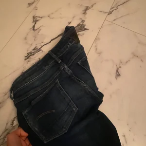 Mörkblå bootcut jeans med råa kanter - Säljer ett par mörkblå lågmidjade jeans med bootcut passform och råa, avklippta benslut. Jeansen har klassiska fem fickor och snygga slitningar framtill. Perfekta för dig som gillar en avslappnad och trendig look.