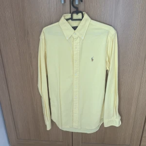 Gul skjorta från Polo Ralph Lauren - Fräsch gul skjorta från Polo Ralph Lauren i slim fit-modell. Klassisk button-down krage, broderad logga på bröstet och långa ärmar. Skjortan är tillverkad i bomull och har knappar framtill. Perfekt för dig som gillar stilrena och färgglada plagg.