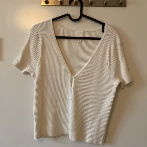 Vit ribbad blus från H&M - Vit ribbad blus från H&M i storlek XL. Toppen har korta ärmar, djup v-ringning och knappar framtill. Materialet är mjukt och stretchigt, perfekt för en chill och stilren look. Enkel att matcha med jeans eller kjol.