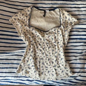 Blommig topp  - Supersöt topp från Divided i ljusbeige med små blå blommor och diskreta ränder. Modellen har korta ärmar, fyrkantig ringning med blå spetskant och figurnära passform. Perfekt för dig som gillar romantiska och feminina detaljer.