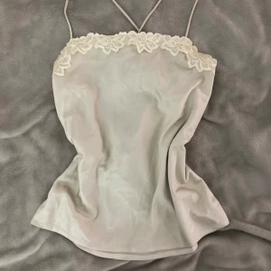 Ljusbeige spetslinne från BikBok - Supersött ljusbeige linne från BikBok med tunna axelband som korsas i ryggen. Toppen har vacker spetsdetalj längs urringningen och är tillverkad i ett glansigt, mjukt material som ger en lyxig känsla. Perfekt för att lyfta vilken outfit som helst. Är i bra skick med sönder i ryggen, de sista bilden