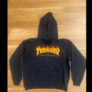 Svart Thrasher hoodie  - Svart hoodie från Thrasher med ikoniskt flames-tryck i gult och orange på bröstet. Tröjan har huva med snörning och en stor magficka. Materialet är 90% bomull och 10% polyester, vilket gör den både mjuk och skön. Perfekt för dig som gillar streetwear och skate.