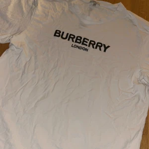 Vit Burberry t-shirt  - Vit t-shirt från Burberry London i 100% bomull. Stor svart logga framtill med klassiskt rutmönster i bokstäverna. Rund halsringning och korta ärmar. Storlek xxl men liten i storleken så skulle säga som en L . Nypris är ca 4000kr