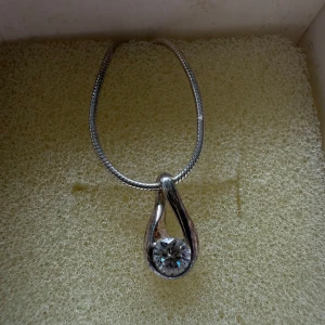 Silverhalsband med droppformad kristall - Ett stilrent silverhalsband med en tunn kedja och ett droppformat hänge med en gnistrande klar kristall. Kommer i en söt vit ask med svart rosett, perfekt som present. Halsbandet har justerbar längd och ger en elegant touch till din look.