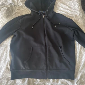 Svart hoodie från Polo Ralph Lauren - Svart hoodie från Polo Ralph Lauren med dragkedja, huva med snörning och klassisk logga på bröstet. Tröjan har två fickor framtill och är tillverkad i mjuk bomullsblandning. Perfekt för en chill och stilren look.