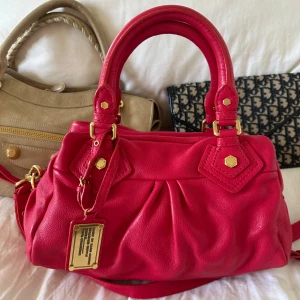 Röd rosa Q Classic Baby Groove väska! - Så fin röd handväska från Marc by Marc Jacobs i äkta läder med guldfärgade detaljer och dubbla handtag. Långa bandet finns med. Otroligt fint skick, som ny. Inget att anmärka på, lädret i toppskick. Klassisk och tidlös design som passar både till vardag och fest. Modellen är rymlig med praktiska fack och dragkedja perfekt för att organisera dina saker. Välbevarad vintage modell som ger en stilren och exklusiv touch till varje outfit. Perfekt till skolstarten eller jobb🤍  Mått: 34x18x12 cm