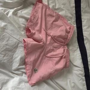 Rosa Moncler shorts med snörning - Snygga ljusrosa shorts från Moncler med elastisk midja och snörning framtill. Klassisk Moncler-logga på benet och mjukt syntetmaterial som känns chill och lätt. Perfekta för sommardagar när du vill ha en clean och fräsch look.