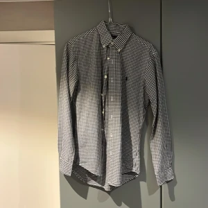 Rutig skjorta från Ralph Lauren - Klassisk svartvit rutig skjorta från Ralph Lauren med button-down krage och broderad logga på bröstet. Skjortan har långa ärmar, vita knappar och är tillverkad i mjuk bomull. Perfekt för en stilren och avslappnad look. Slimfit
