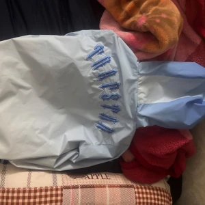 Ljusblå vindjacka från trapstar - Snygg ljusblå vindjacka från trapstar  med huva, dragkedja och coolt broderat märke på ärmen. Jackan har tryckknappar på fickorna och broderad text på ryggen. Perfekt för dig som vill sticka ut med stilren streetwear.