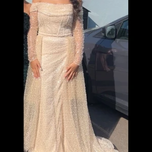 Champagnefärgad glittrig festklänning  -  långklänning i champagnefärg med glittriga paljetter. Offshoulder-modell med transparenta långa ärmar. Beige förlovningskläning köpt för 17 000 kr. Den är i bra skick och använd 1 gång. Den har även ett släp till som går att sätta på och ta av. Kläningen är inte så tung som den ser ut och vara vilket är skönt för att kunna röra sig med den. Klänningen har även små pärlor vilket gör den så unik. Eftersom kläningen har band som knyts som korset så passar den i nästan alla storlekar från XS-M .