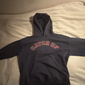 Mörkblå hoodie Pull&Bear CATCH UP - Mörkblå hoodie från Pull&Bear med stor huva och magficka. På bröstet står det 'CATCH UP' i röda bokstäver med vit kant. Perfekt för chill dagar och enkel att matcha med jeans eller joggers.