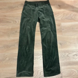 Mörkgröna Juicy Couture byxor - Mörkgröna Juicy Couture del ray byxor med bred resår och snörning i midjan. Byxorna har raka ben, stora fickor fram och bak med knappdetaljer. Perfekta för en chill och trendig look.