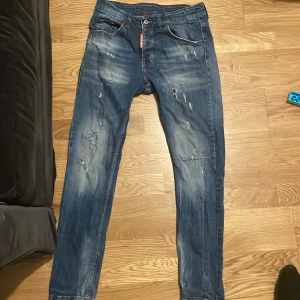 jeans från Dsquared2 - Säljer ett par blå jeans från Dsquared2 med coola slitningar och tvättade detaljer. Klassisk femficksmodell med låg midja och smal passform. Ikonisk Dsquared2-lapp bak i grönt och vitt samt röd logga vid fickan. Perfekta för en trendig streetstyle.