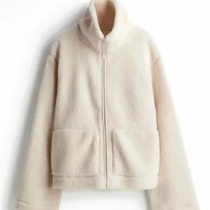 Beige teddyjacka med dragkedja - Mysig beige teddyjacka med hög krage och två stora fickor framtill. Jackan har hel dragkedja och är tillverkad i fluffigt teddy-material som ger en chill och trendig look. Perfekt för lager-på-lager-stil under kyliga dagar.