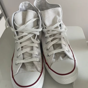 Vita Converse All Star high tops - Klassiska vita Converse All Star high tops med röd och blå rand runt sulan. Skorna har snörning, rund tå och är gjorda i canvas med gummisula. Priset kan diskuteras.