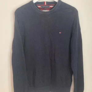 Mörkblå stickad tröja Tommy Hilfiger - Mörkblå tröja från Tommy Hilfiger i pima bomullsblandning med kashmir. Klassisk rund halsringning med ribbade muddar och broderad logga på bröstet. Perfekt för dig som gillar stilrena och bekväma plagg.