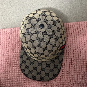 Gucci keps med GG-mönster - Snygg keps från Gucci i beige och svart med klassiskt GG-monogram över hela kepsen. Kepsen har en böjd skärm och en röd-blå rand på sidan. Tillverkad i bomullscanvas med mörkblått innerfoder och justerbar rem bak.