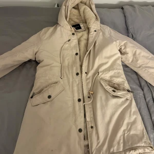 Beige parkasjacka med teddyfoder Copperose - Snygg beige parkasjacka från Copperose med mjukt teddyfoder och huva. Jackan har dragkedja, knappar, snörning i midjan och stora fickor med dragkedja. Perfekt för kalla dagar när du vill vara både varm och stilren.