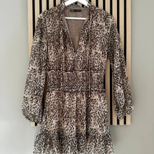 Leopardmönstrad kortklänning från Zara - Säljer en snygg kortklänning från Zara i leopardmönster med v-ringning, smockad midja och volang nertill. Klänningen har långa, lätt puffade ärmar och är tillverkad i ett tunt, luftigt material som känns superbekvämt. 💕 Endast använd en gång. 