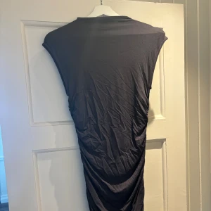 Svart medellång klänning från Gina Tricot - Svart medellång klänning i från Gina Tricot i mjuk bomull. Klänningen har ärmlös design, rund hals och snygg rynkdetalj längs ena sidan som ger en cool omlott-effekt. 