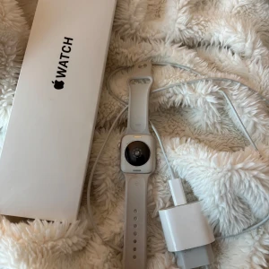 Apple Watch - Apple Watch i silver med vit sportrem, komplett med originalförpackning och laddare. Klockan ser ut att vara i gott skick utan synliga repor eller skador. Perfekt för träning, hälsa och smarta notiser direkt på handleden.