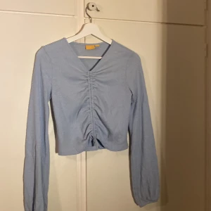 Ljusblå ribbad topp från Gina Tricot - Supersöt ljusblå långärmad topp från Gina Tricot med ribbad struktur och snygg rynkdetalj framtill. Toppen har v-ringning och croppad passform, perfekt till jeans eller kjol. Materialet är mjukt och stretchigt för en bekväm känsla.