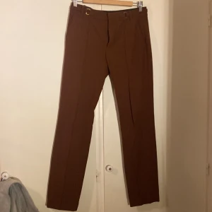 Bruna kostymbyxor från Zara Woman - Snygga bruna kostymbyxor från Zara Woman med raka ben och pressveck. Byxorna har hög midja, bälteshällor med guldfärgade detaljer och diskreta fickor bak. Materialet är en blandning av polyester och viskos som ger en dressad känsla. Perfekt för dig som gillar att vara uppklädd under en vardag.