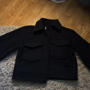 Marinblå kort kappa från Gina Tricot - Marinblå kort kappa från Gina Tricot med två stora bröstfickor och klassisk krage. Jackan är croppad och har en rak passform med knäppning framtill. Materialet är mjukt och påminner om ull, perfekt för lager-på-lager-stil.