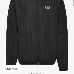Svart vindjacka från EA7 Emporio Armani - Svart vindjacka från EA7 Emporio Armani med vit logga på bröstet. Jackan har huva, dragkedja framtill och två sidofickor med dragkedja. Tillverkad i lätt polyester, perfekt för blåsiga dagar. Enkel och stilren design som funkar till det mesta.