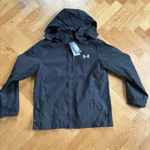 Svart vindjacka från Under Armour - Svart vindjacka från Under Armour med huva och dragkedja framtill. Jackan har två sidofickor med dragkedja och meshfoder på insidan för extra ventilation. Snyggt logotryck på bröstet och justerbara detaljer vid huvan. Perfekt för blåsiga dagar.